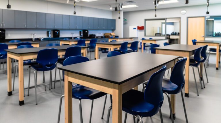 Modern science lab classroom with black work surfaces and blue chairs arranged at lab tables, featuring built-in cabinetry, sinks, and ceiling-mounted utilities for hands-on experiments.