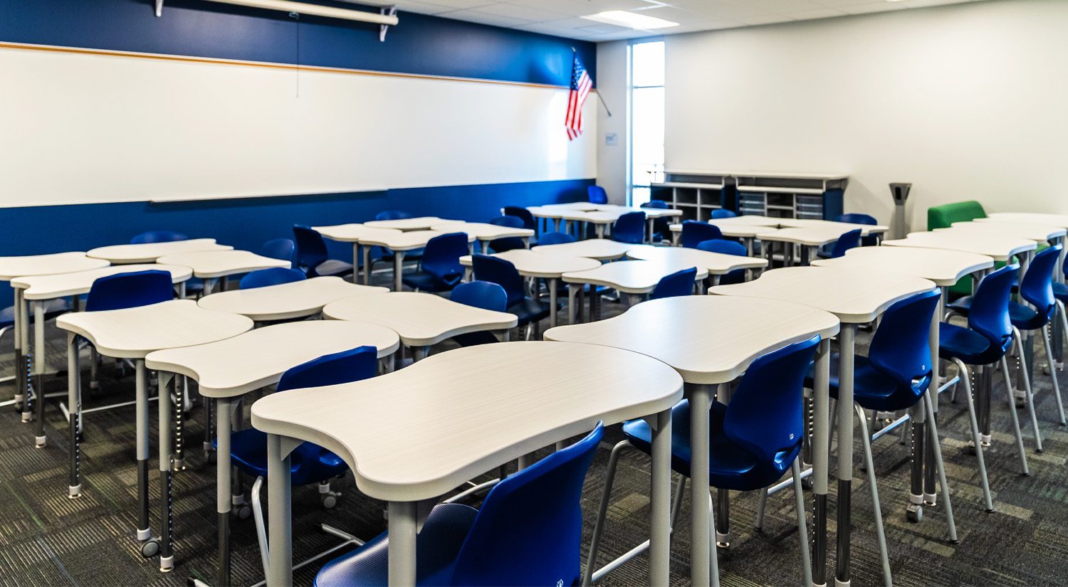 mpty modern classroom with flexible, curved desks and blue chairs arranged for collaboration, featuring a whiteboard wall, storage cubbies, and natural light from a window.