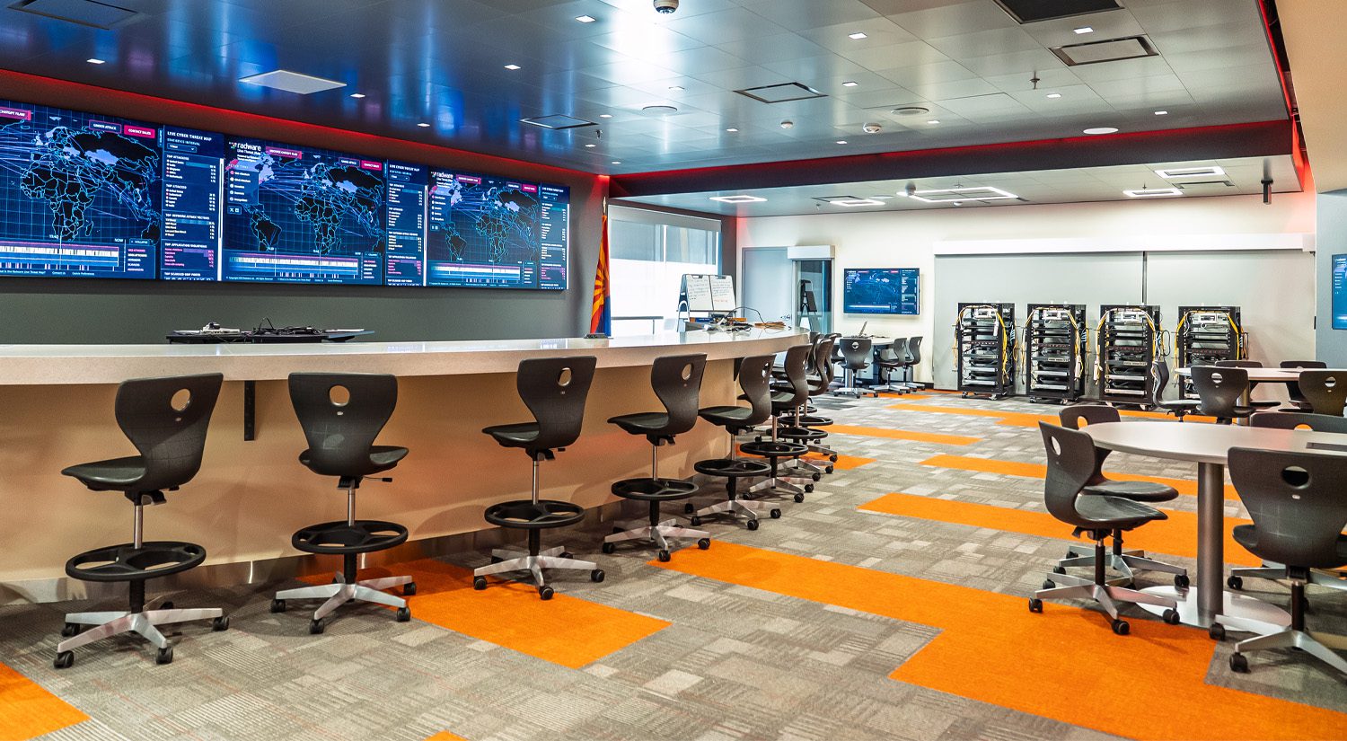 Modern technology classroom or cybersecurity lab with high stools and collaborative tables, featuring a wall of large digital monitoring screens, server racks, and a front counter workspace designed for hands-on, real-world learning.