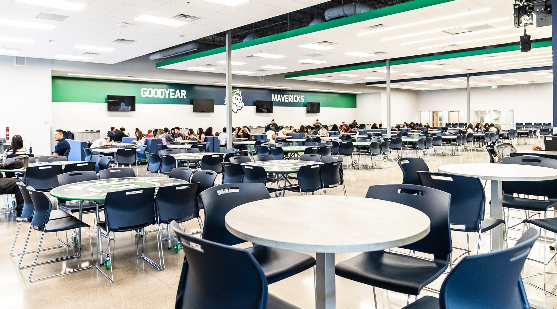 Spacious high school cafeteria with round tables and blue chairs, where students gather and collaborate under bright lighting, with school branding displayed on the walls.