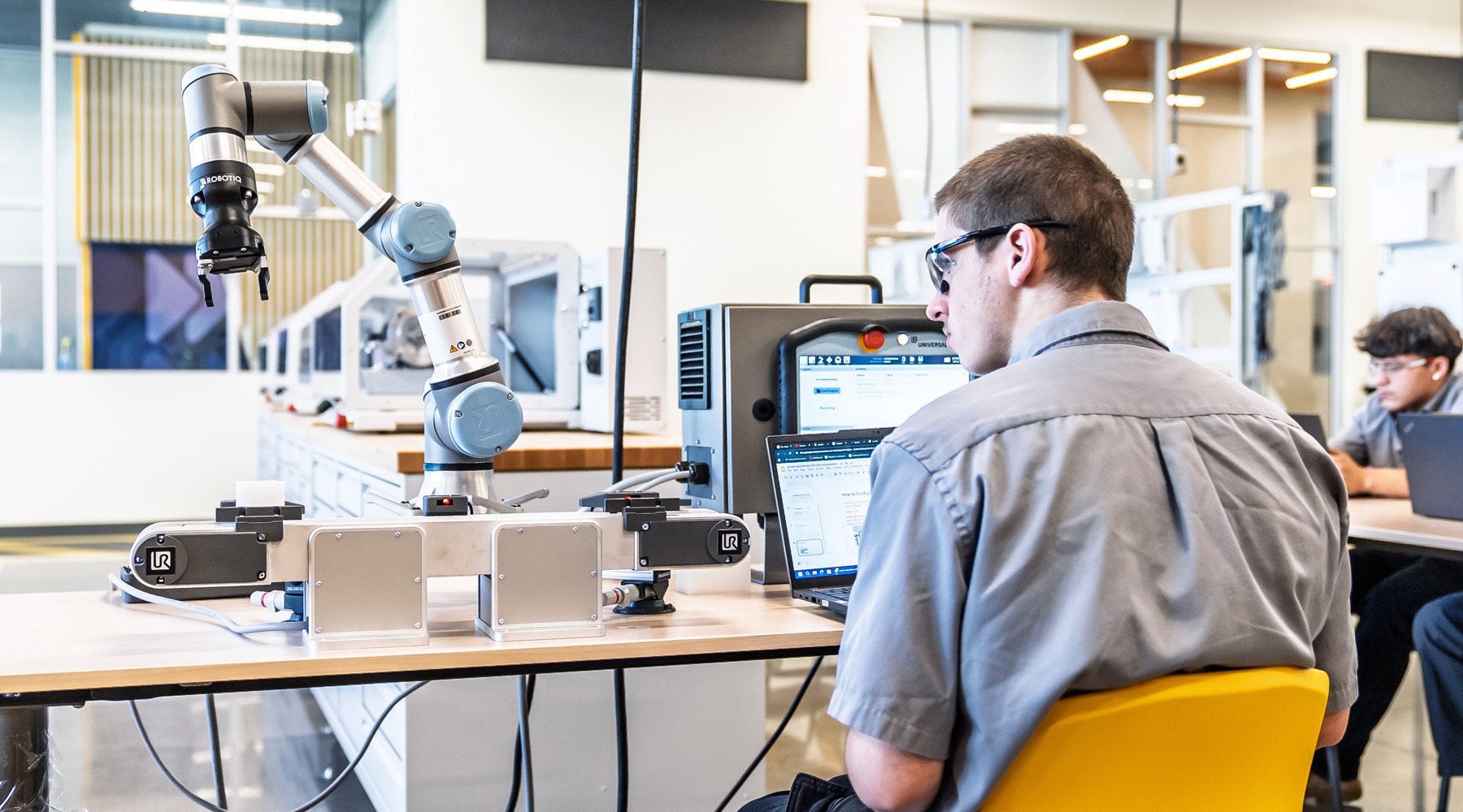 Student working on a laptop programs a robotic arm in a modern robotics lab, with advanced equipment and peers collaborating in the background.