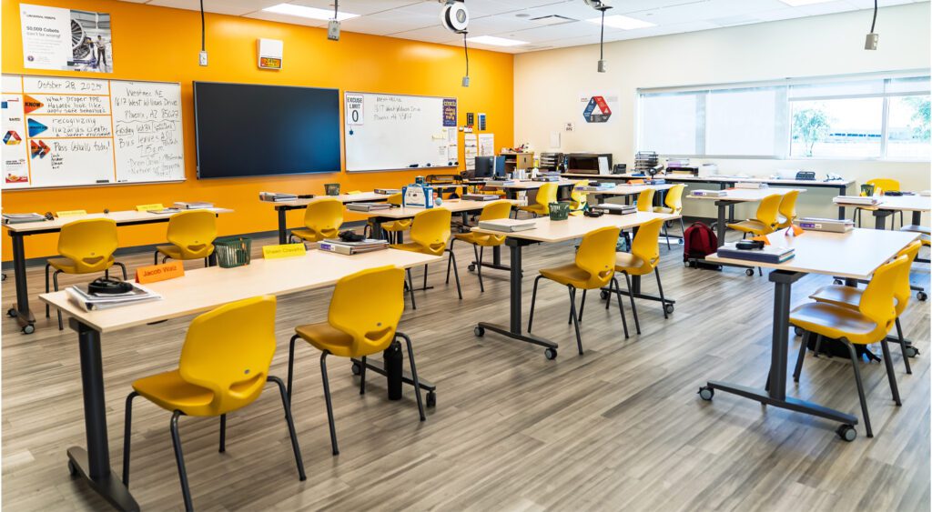 Bright, modern classroom with yellow chairs and mobile desks arranged for group learning, featuring a large digital display, whiteboards with lesson notes, ceiling-mounted power reels, and natural light from wide windows.