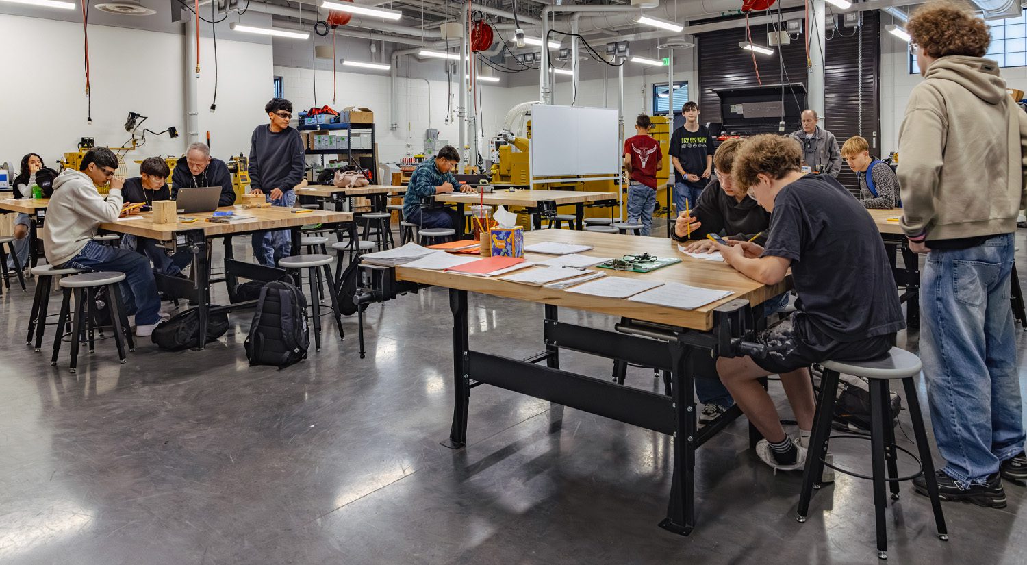 Students collaborate and work independently at tables in a modern high school CTE lab classroom equipped with tools, machinery, and flexible workspaces, while a teacher assists nearby.