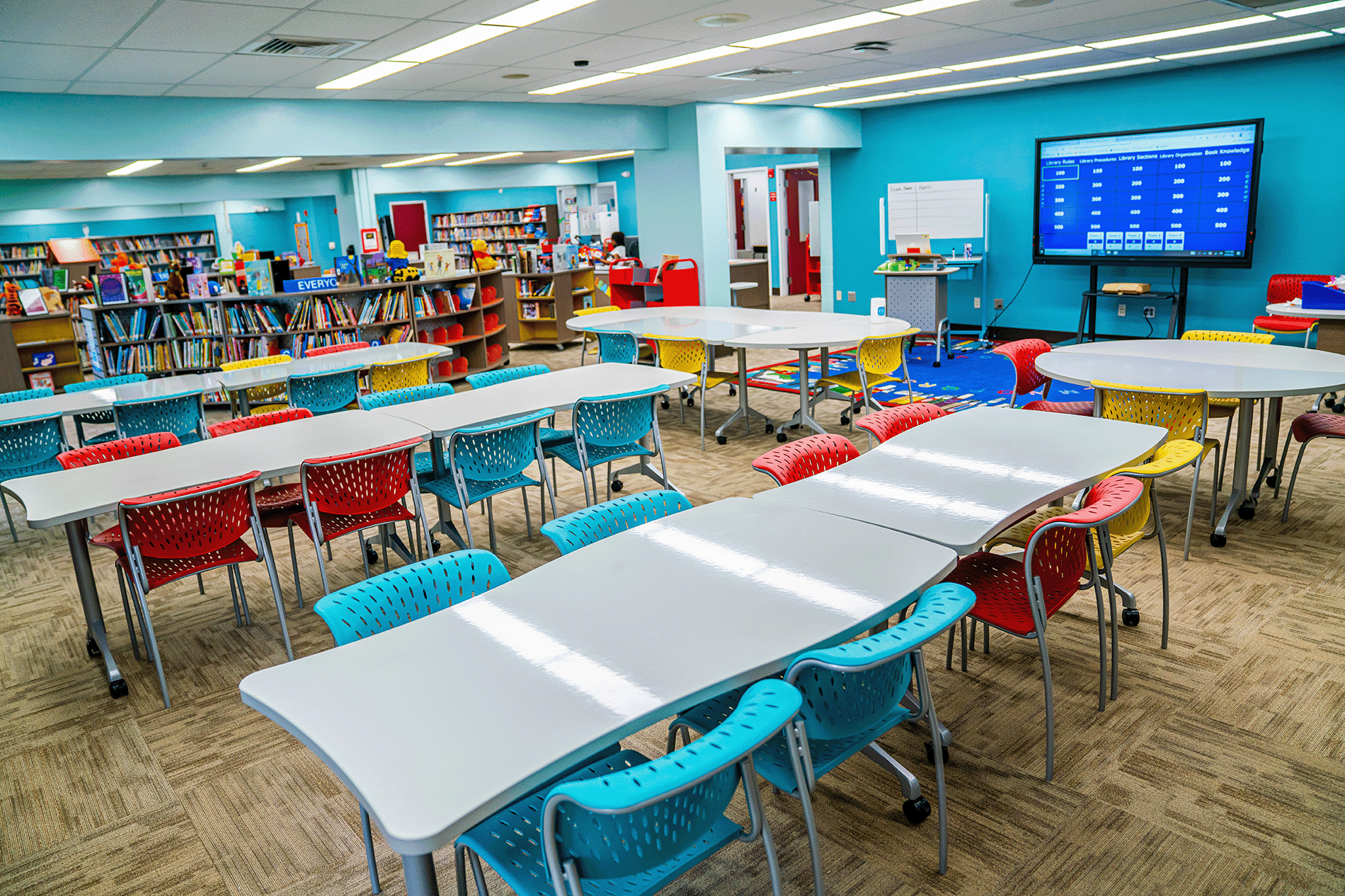Bright and colorful school media center featuring flexible white tables, red, blue, and yellow chairs, bookshelves filled with books, and a large interactive display screen.