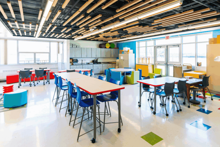 Colorful and modern classroom with various flexible seating options including blue and red high-top stools, black chairs at wheeled tables, and soft seating in green, blue, and yellow. The space features bright natural light from large windows, ample storage with gray cabinetry, and an artistic wood slat ceiling with overhead lighting. The classroom supports collaborative learning and creative activities.