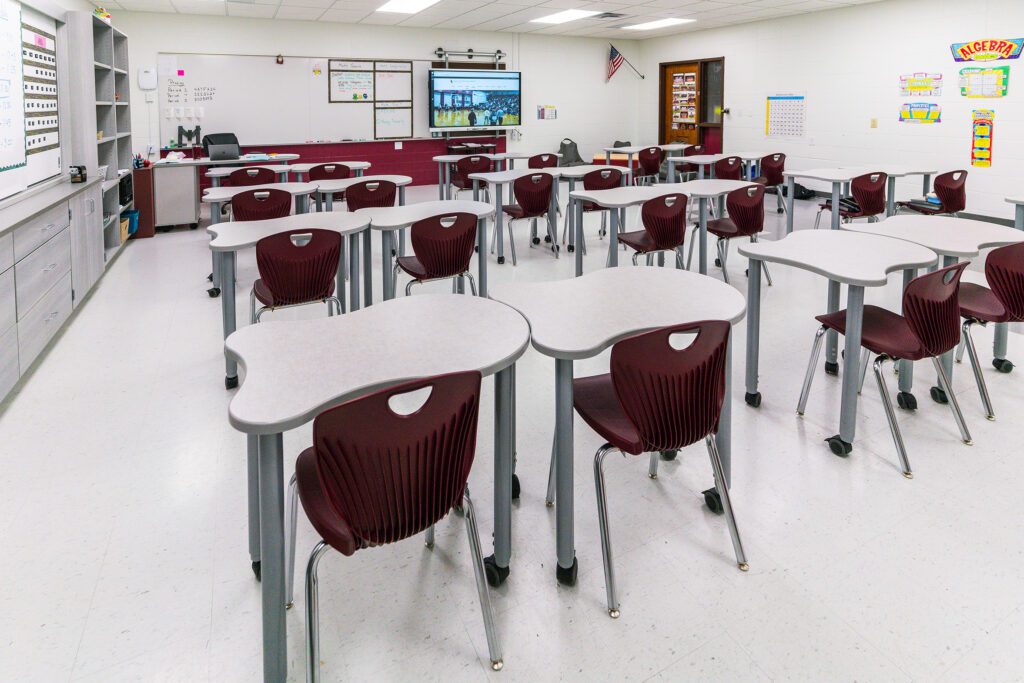 Modern classroom with individual student desks and maroon chairs arranged in rows, facing a smartboard and whiteboard at the front of the room. Educational posters are displayed on the walls.