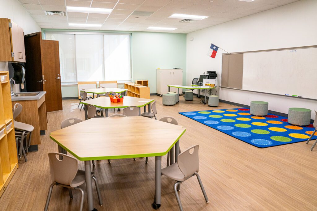 Early childhood classroom with hexagon tables, soft seating, colorful carpet, and a Texas flag above the whiteboard.
