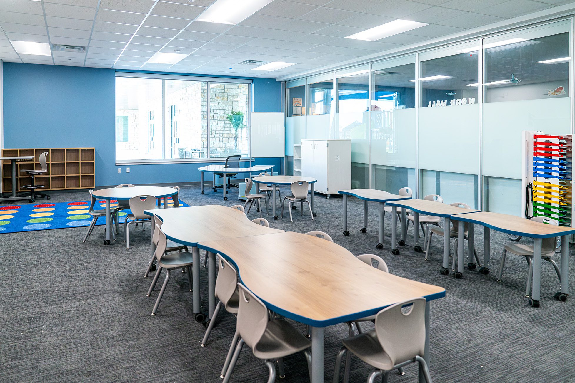 Elementary classroom with curved tables, neutral chairs, and colorful carpet, featuring large windows and open shelving.