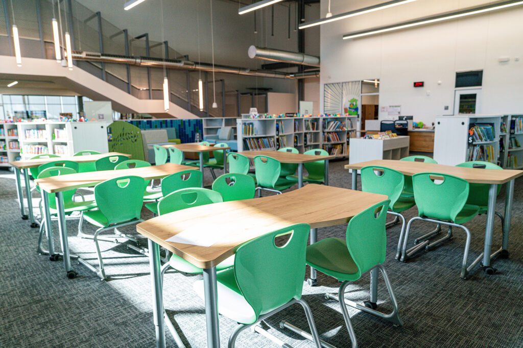 Modern school library with green chairs, wood-top desks, open book shelving, and natural lighting.