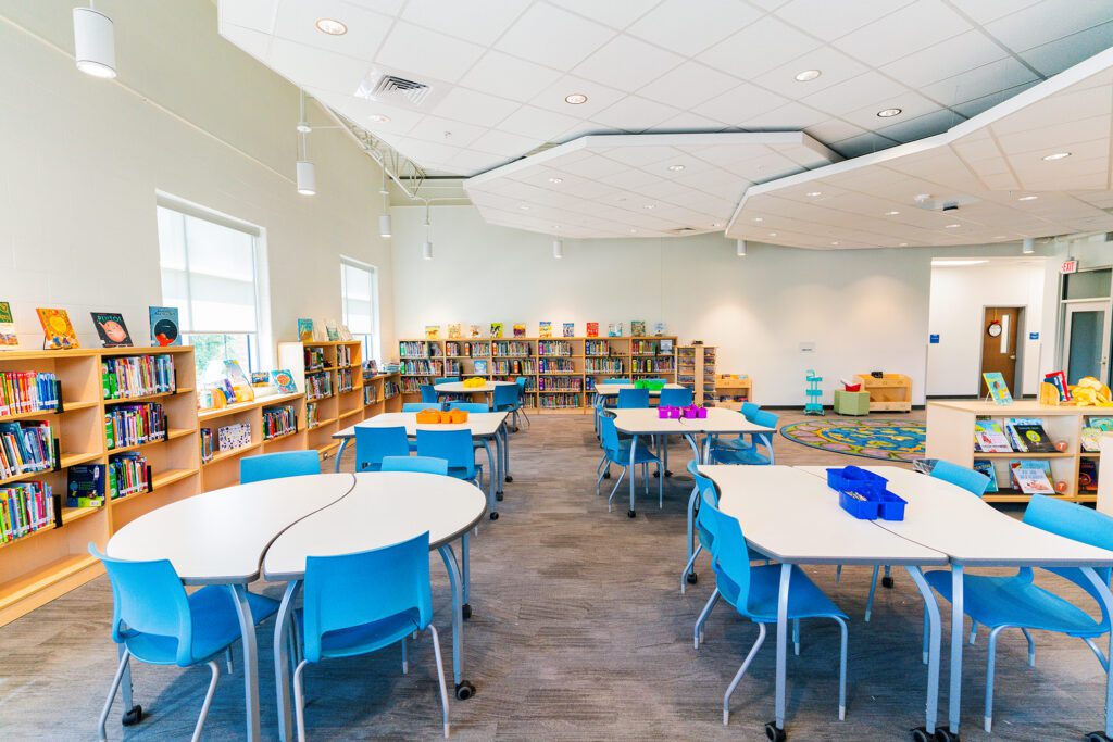 Bright elementary school library with low bookshelves, colorful children’s books, round collaborative tables, and blue chairs in an open, flexible learning space.