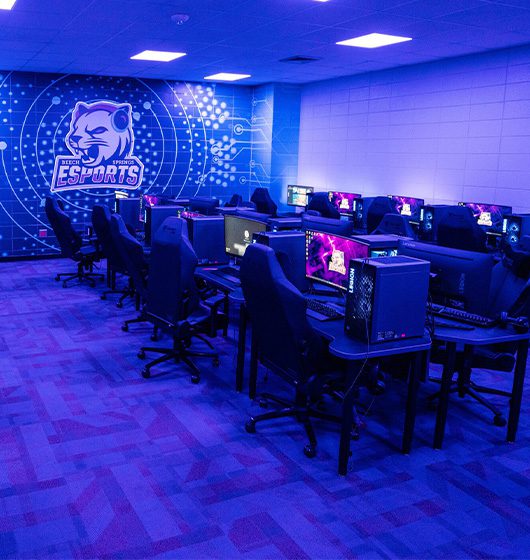 Modern middle school esports lab with gaming PCs, blue lighting, and a team logo on the back wall.