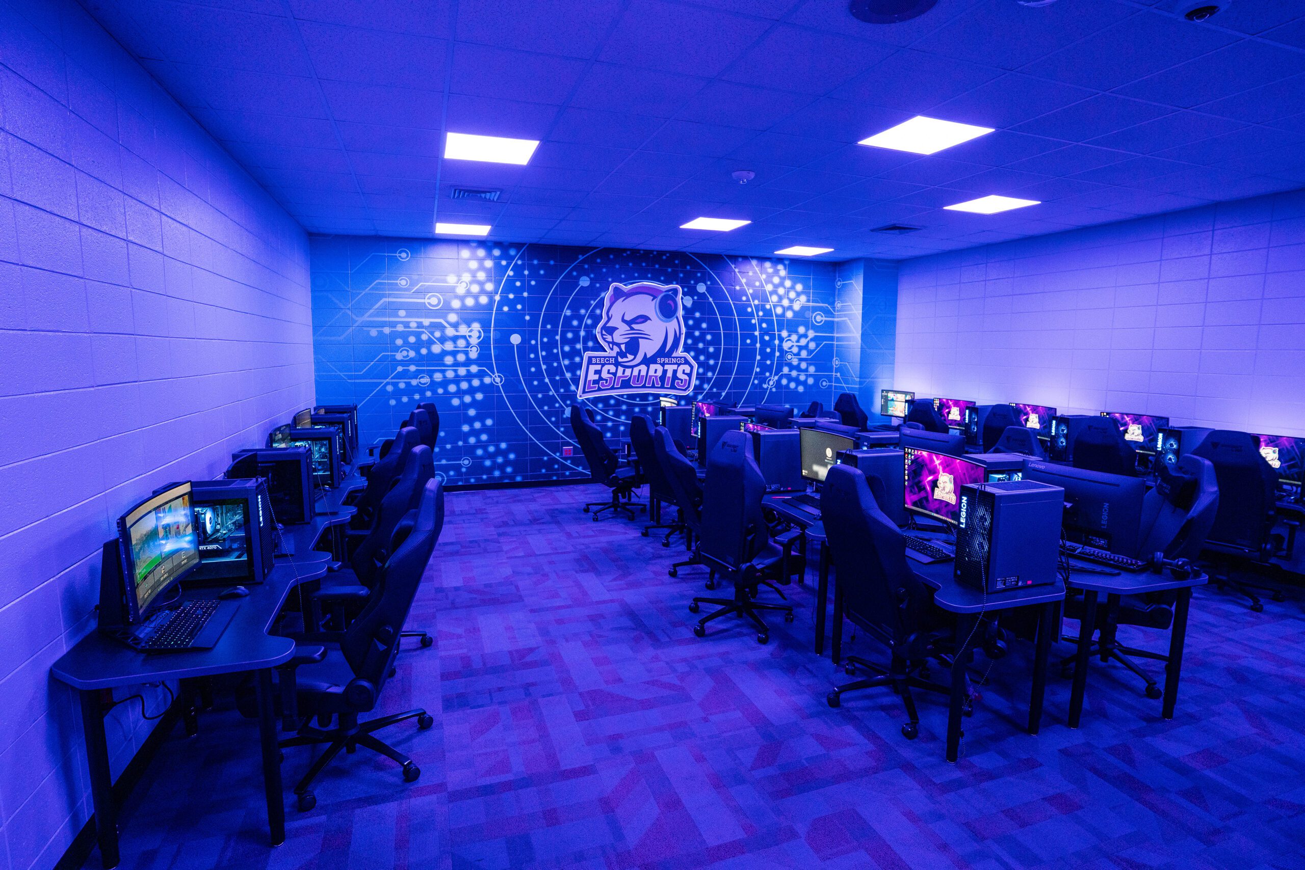Modern middle school esports lab with gaming PCs, blue lighting, and a team logo on the back wall.