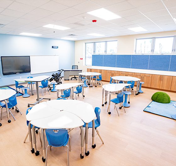 Elementary classroom with round collaborative tables, blue student chairs, wall-mounted display, and built-in cabinetry in a bright, flexible learning space.