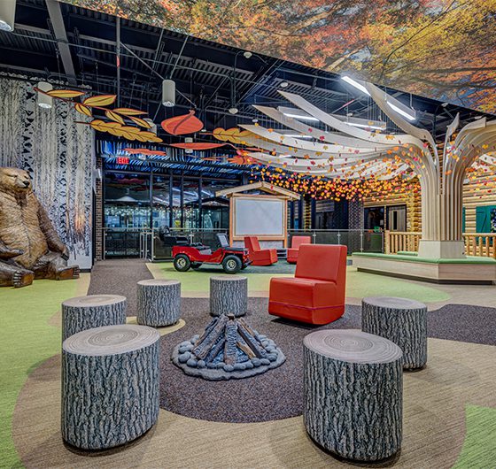 Creative indoor learning commons with treehouse structure, campfire seating area, red lounge chairs, decorative ceiling elements, and flexible collaboration spaces.