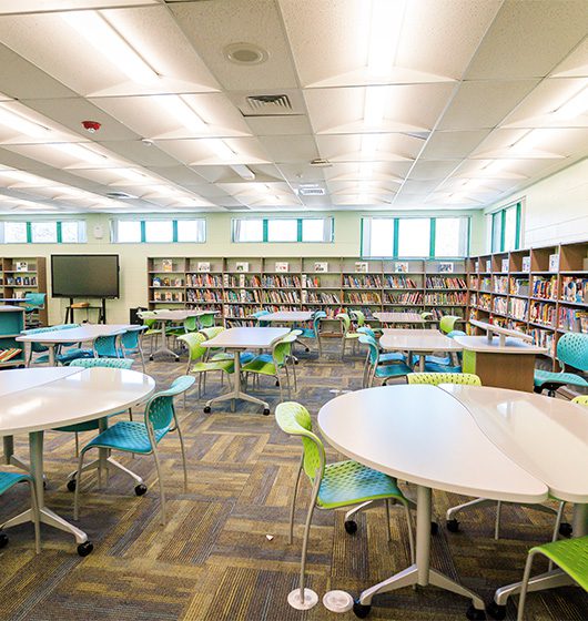 Modern school library with flexible tables, blue and green student chairs, and wall-to-wall bookshelves designed for collaborative learning and reading.