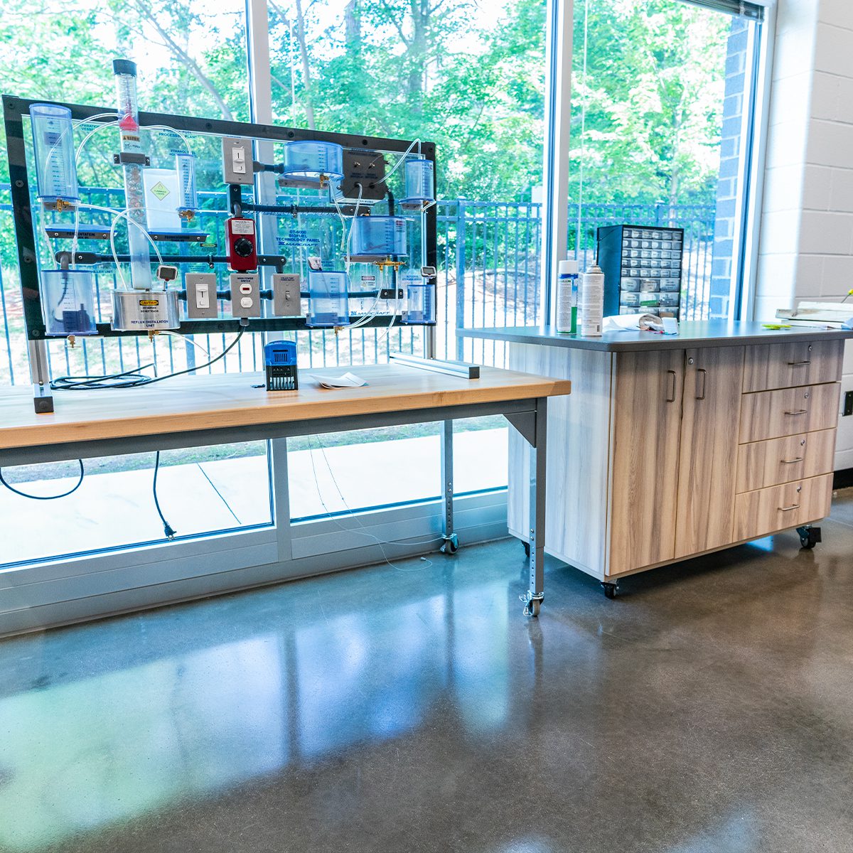 STEM lab workstation with engineering training equipment mounted on a panel, alongside mobile storage cabinets and work tables in a modern classroom with large windows.