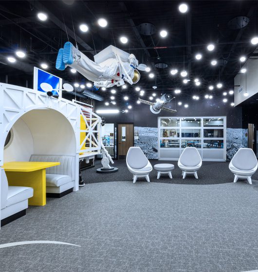 Space-themed collaborative learning environment with astronaut and satellite installations, modern lounge seating, and interactive learning spaces designed to inspire STEM exploration and student engagement.