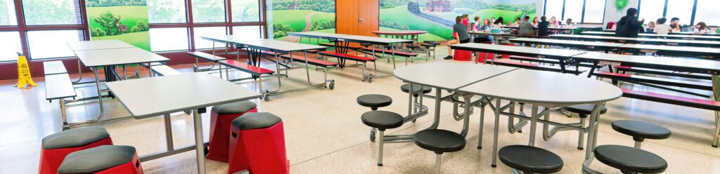 A bright school cafeteria with large windows, mobile folding tables, built-in and freestanding stools in red and black, and students dining in the background alongside a colorful landscape mural on the wall.