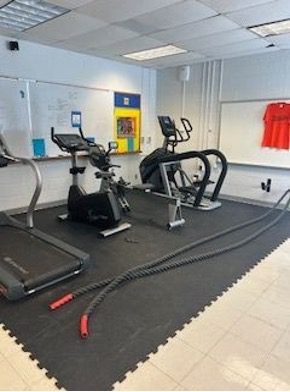 Fitness room with treadmills, a stationary bike, and elliptical machines on black rubber flooring, with battle ropes stretched across the floor.