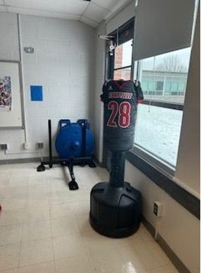 Freestanding boxing punching bag with a red practice jersey pulled over it, positioned near classroom windows in a school fitness space.