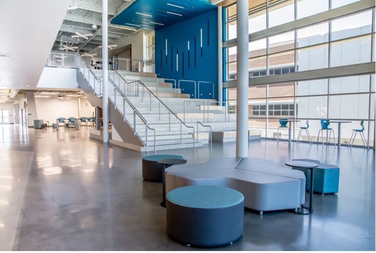 Two-story school atrium with wide tiered seating steps, a blue accent wall with vertical light details, and modern lounge seating below.