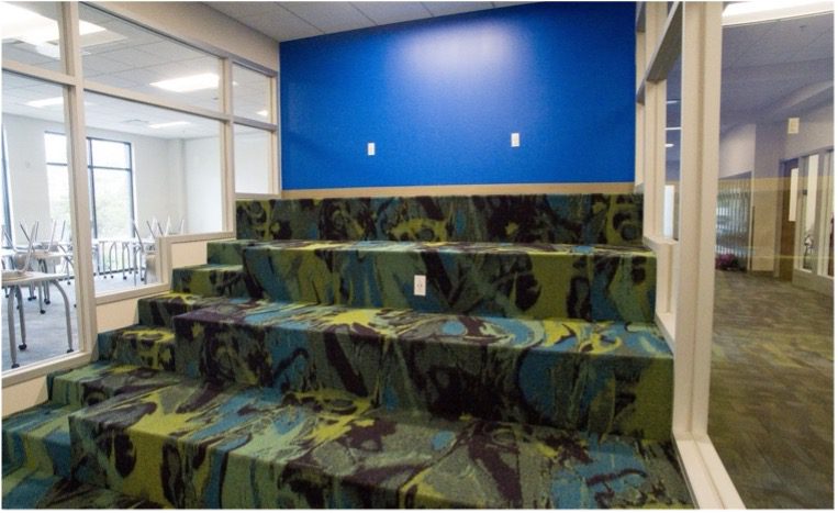 Colorful tiered carpeted steps in blue, green, and purple against a bright blue wall, used as flexible seating between classroom spaces.