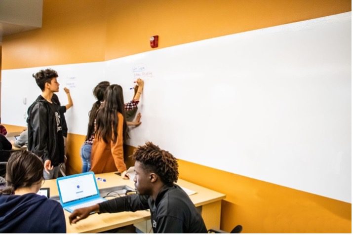 Students writing on a wall-mounted whiteboard together while a classmate works on a laptop at a nearby desk.