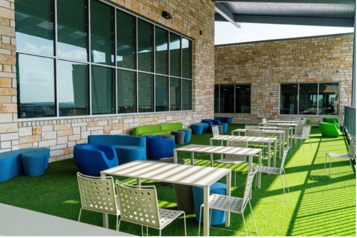 Outdoor learning patio with turf flooring, blue and green lounge chairs, and small tables set against a stone building exterior.